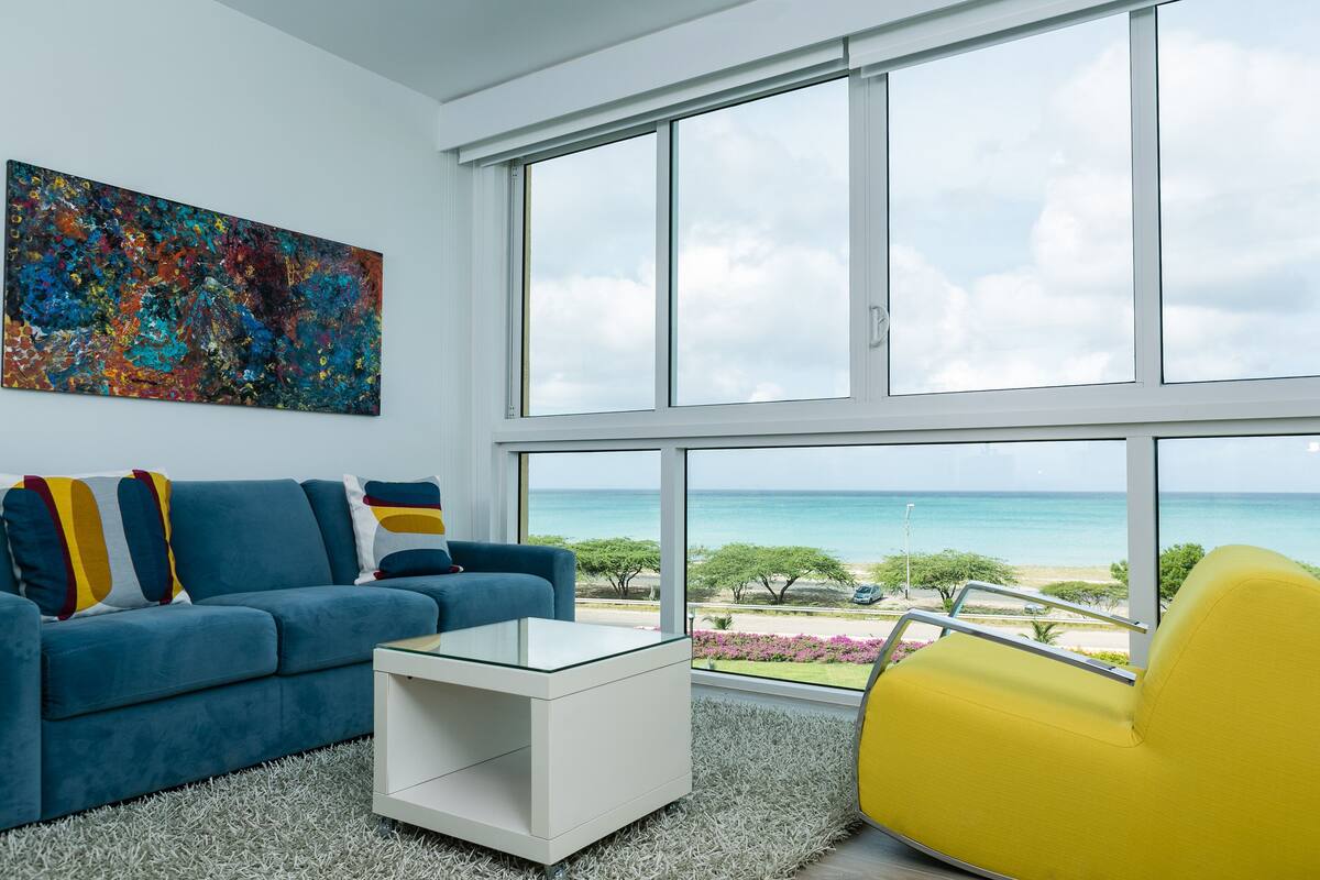 A modern living room with a large window, a blue sofa, a yellow armchair, and a white coffee table.