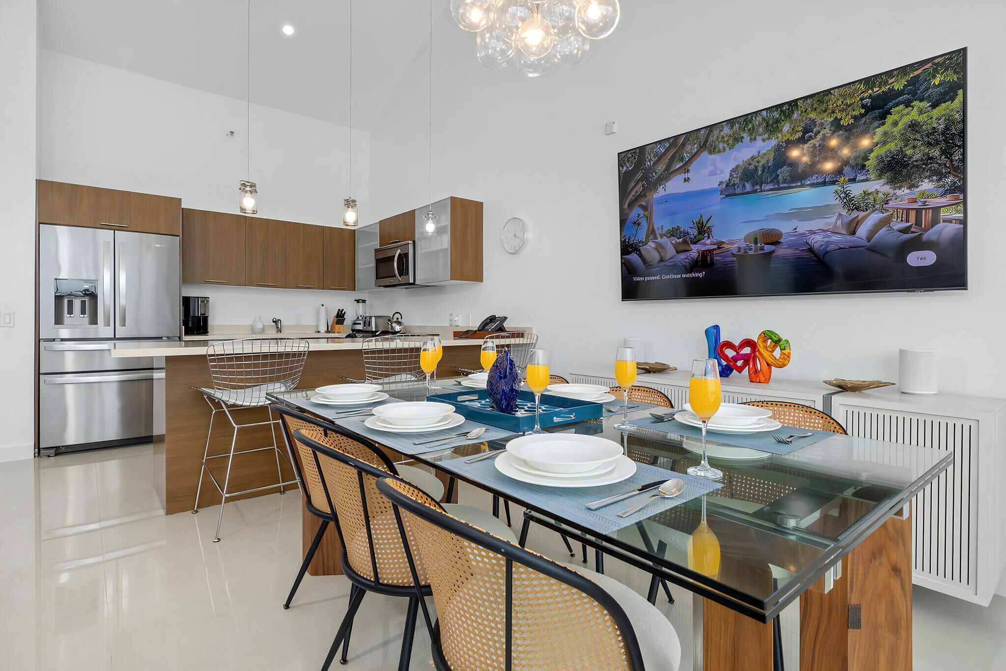 A modern kitchen with stainless steel appliances, a dining area with a glass table, and a large TV displaying a tropical scene.
