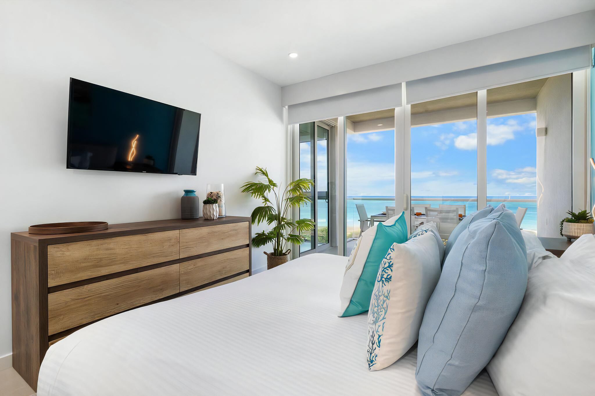 A modern bedroom with a large bed, a TV mounted on the wall, and a view of the ocean.