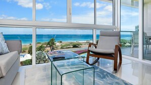 A modern living room with a glass coffee table, a white sofa, and a wooden chair. A large window offers a view of the ocean and beach.