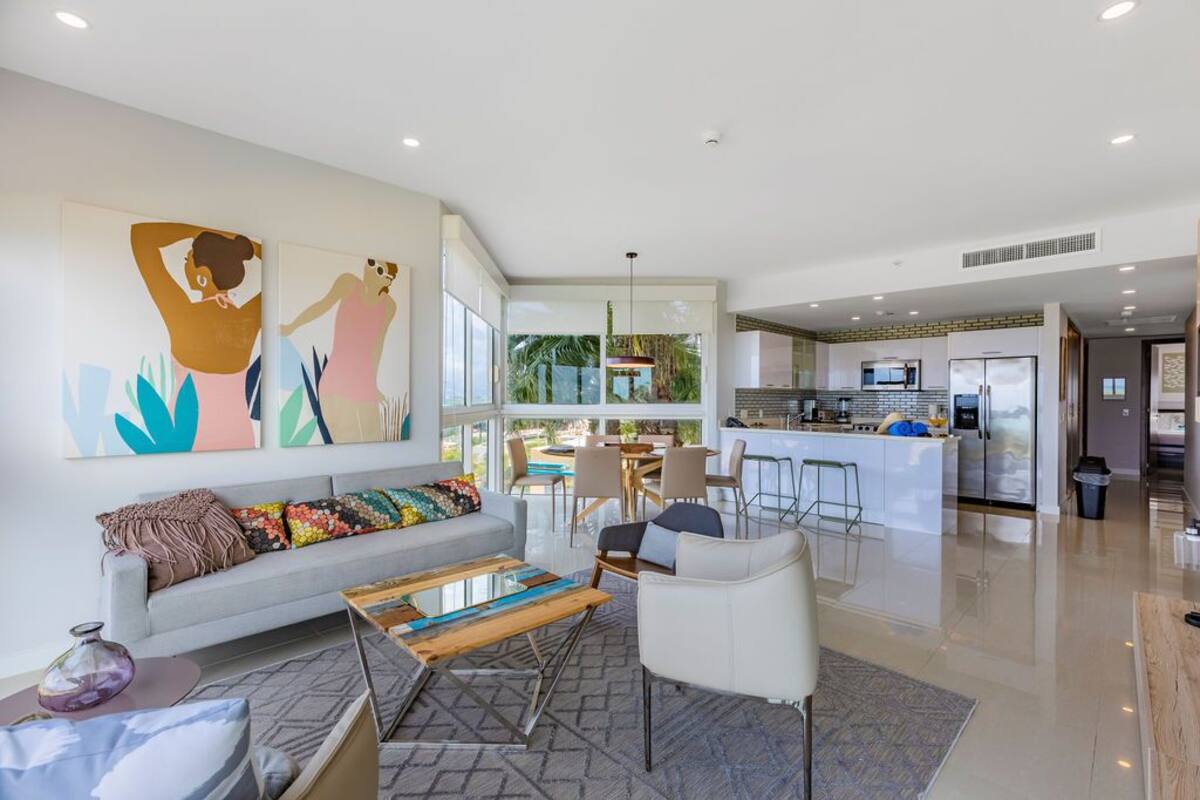 A modern living room with a sofa, coffee table, and artwork. A kitchen with a bar and dining area is visible in the background.