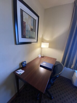 A hotel room with a wooden desk, a framed picture on the wall, a lamp, and a chair.