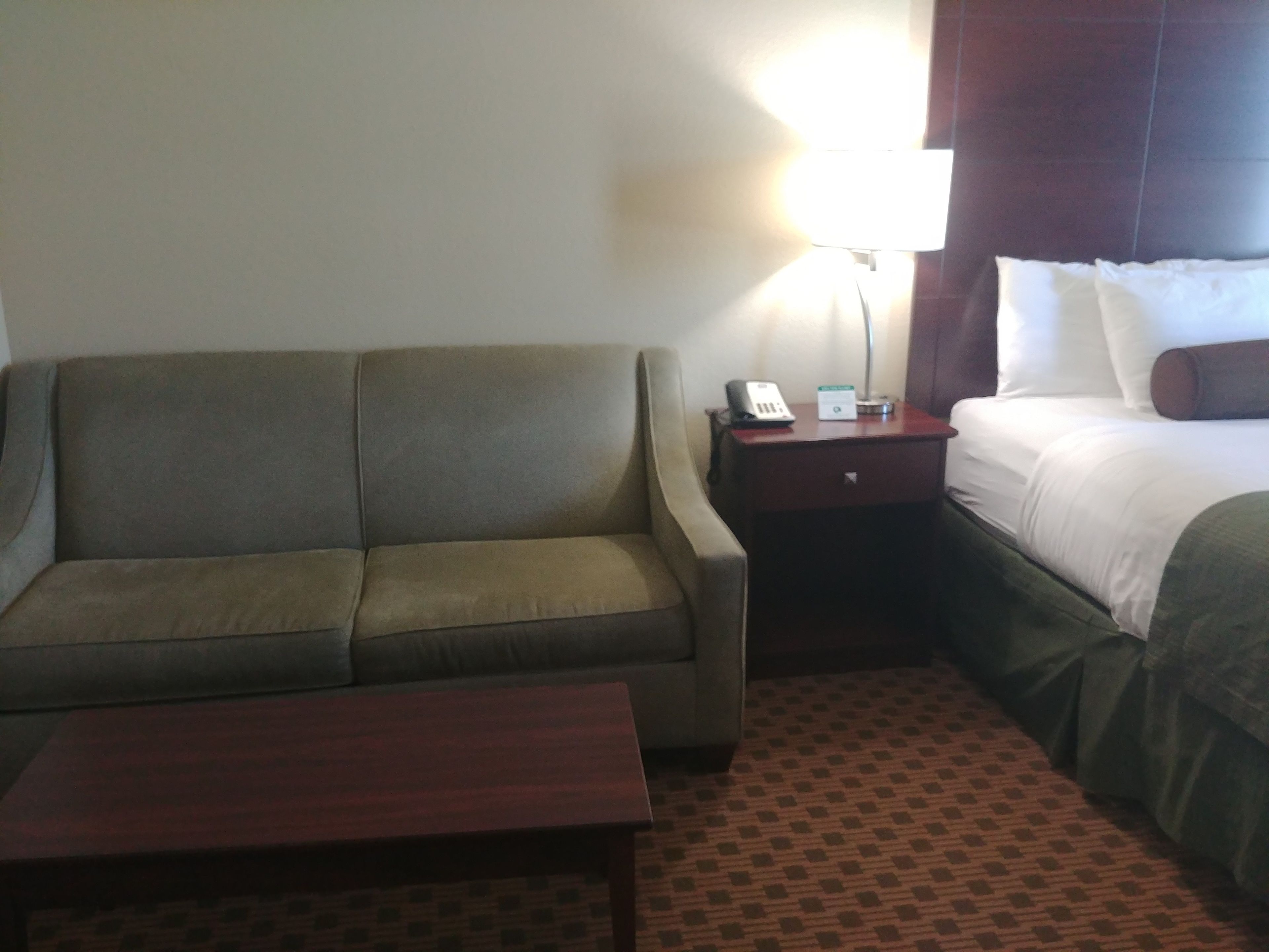A hotel room with a sofa, a bedside table with a lamp, a bed with pillows, and a patterned carpet.