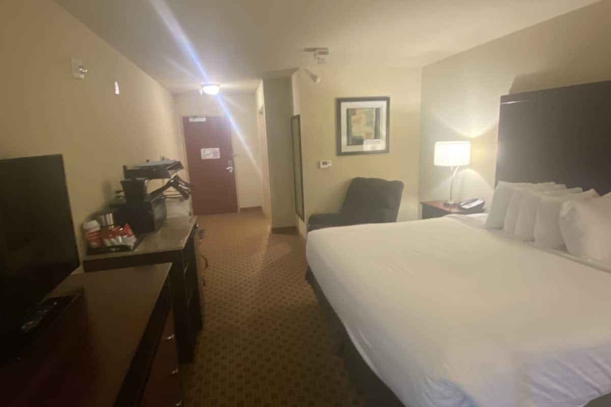 Hotelkamer met een bed, een bureau met een televisie, een stoel, een nachtkastje met een lamp en een deur naar een andere kamer.