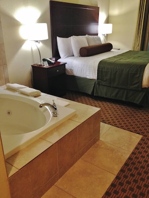 Habitación de hotel con una bañera grande, una cama bien tendida, una mesita de noche con lámpara y un teléfono de pared.