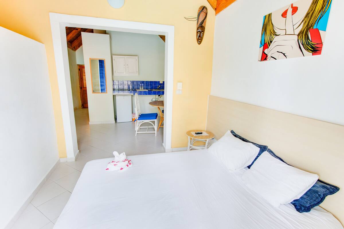 A hotel room with a bed, a small table, and a chair. There is a kitchenette in the background.