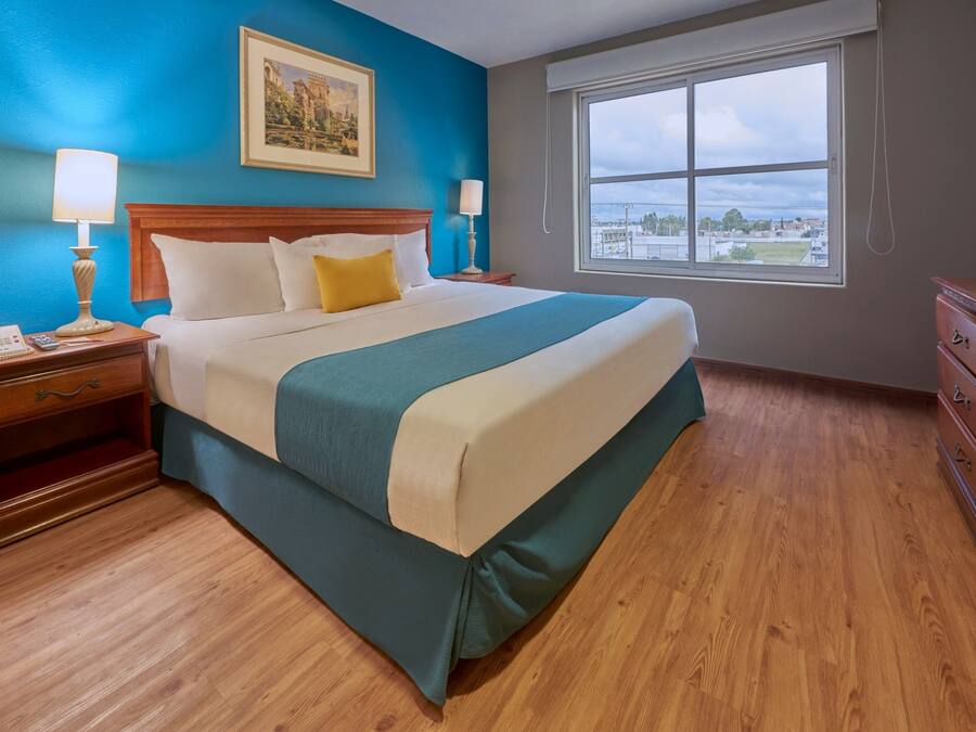 Une chambre d’hôtel avec un grand lit, des tables de chevet en bois, une commode, une fenêtre donnant sur la ville et un tableau au mur.