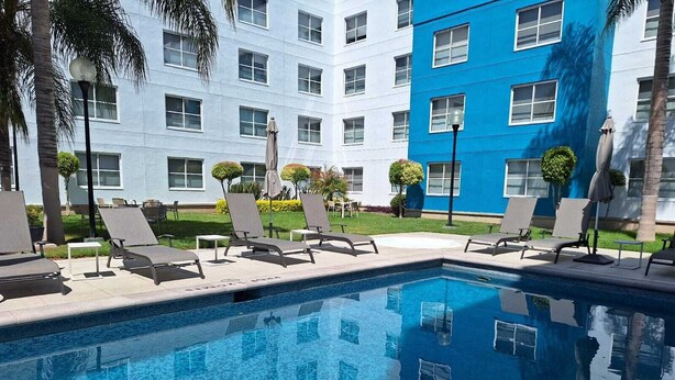 A swimming pool with lounge chairs and umbrellas, surrounded by a building with multiple windows and a blue facade. - Hotel Sierra by HP Hotels (San Luis Potosi)