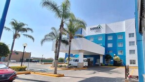 A hotel with a modern design, featuring a prominent blue and white facade, a parking area with several vehicles, and palm trees in the foreground. - Hotel Sierra by HP Hotels (San Luis Potosi)