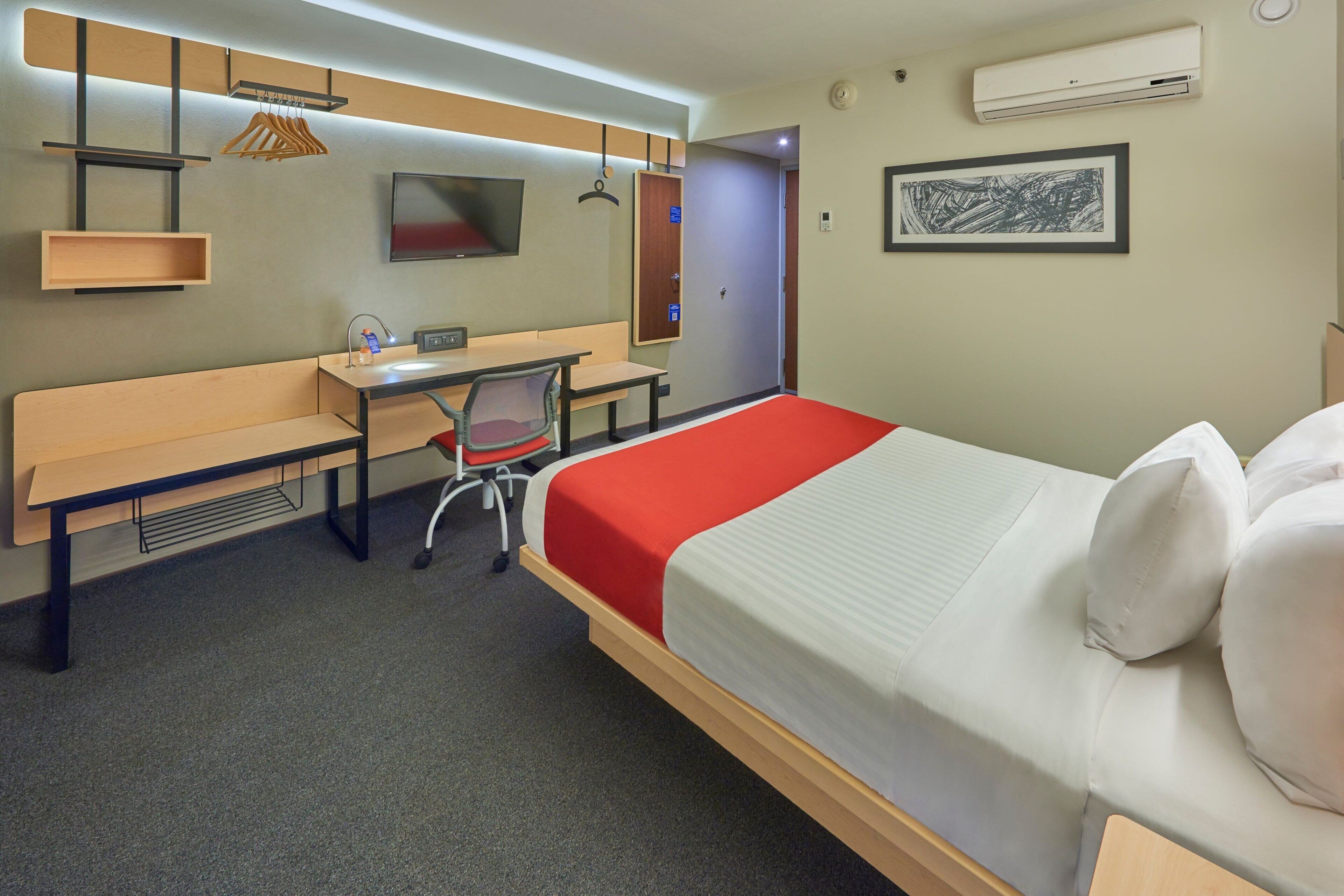 A hotel room with a bed, a desk, a chair, a bench, and a TV.