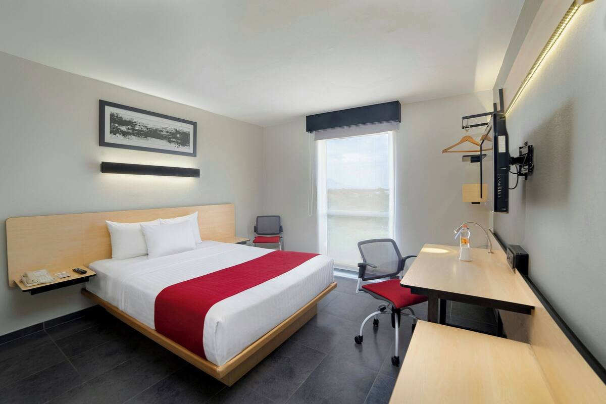 Een hotelkamer met een groot bed, een bureau, een stoel, een klein tafeltje, een nachtkastje, een telefoon, een schilderij aan de muur, een raam en een hanglamp.