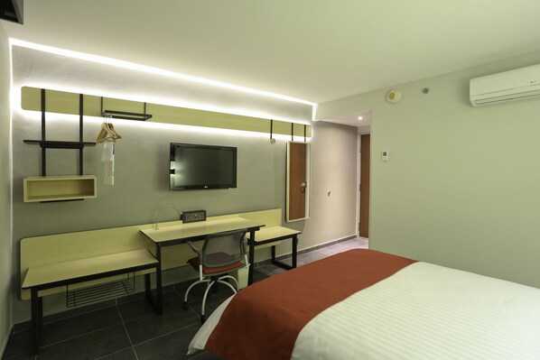 Una habitación de hotel con una cama, un escritorio con silla, un televisor, un espejo y una estantería.