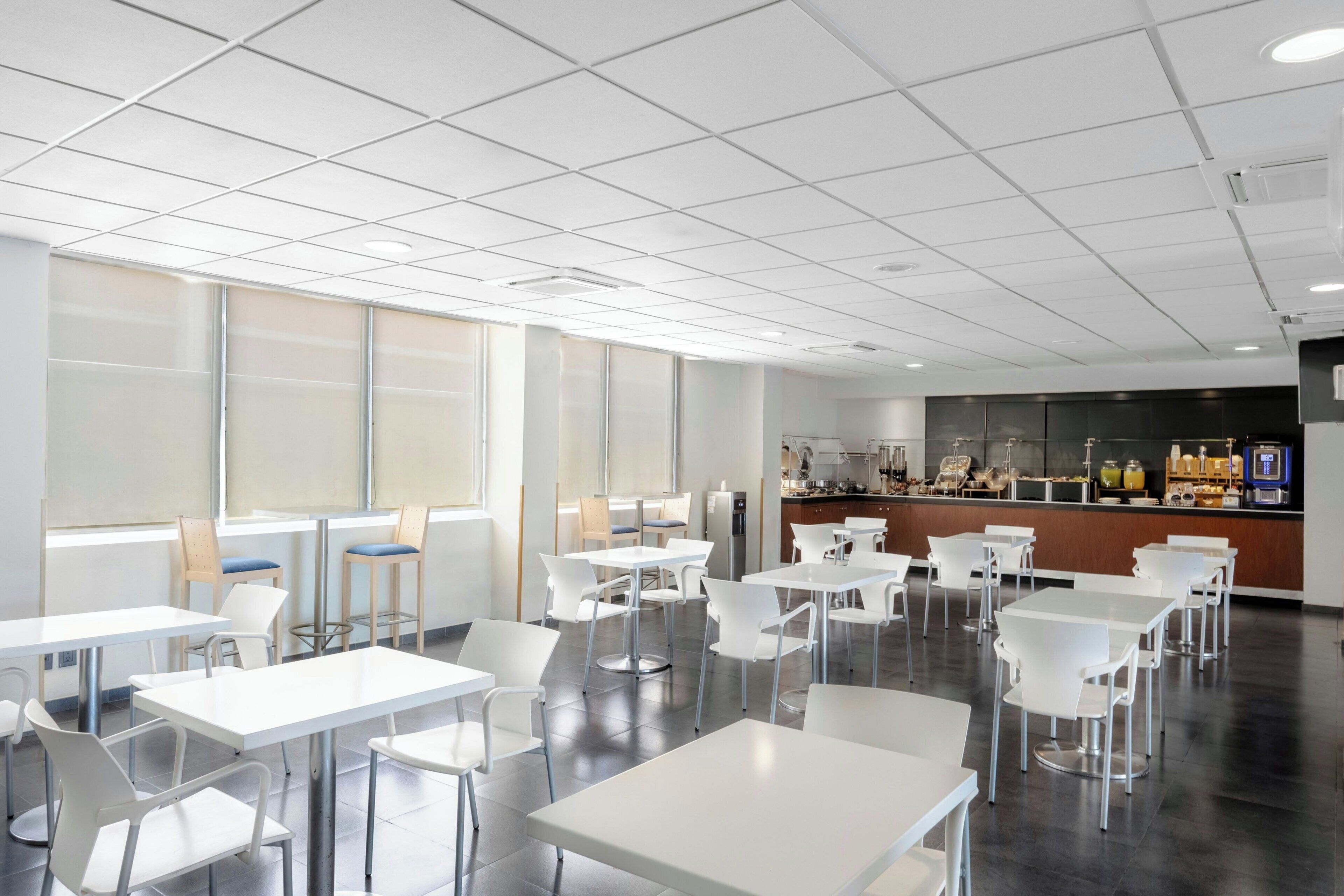 Un espace repas moderne avec des tables et des chaises blanches, un comptoir où sont présentés des plats, et de grandes fenêtres.