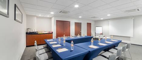Un salón de conferencias con una larga mesa, sillas, una pantalla de proyector y una pequeña área de cocina.