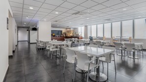 En moderne kantine med hvide borde og stole, et tekøkken og store vinduer der giver naturligt lys.