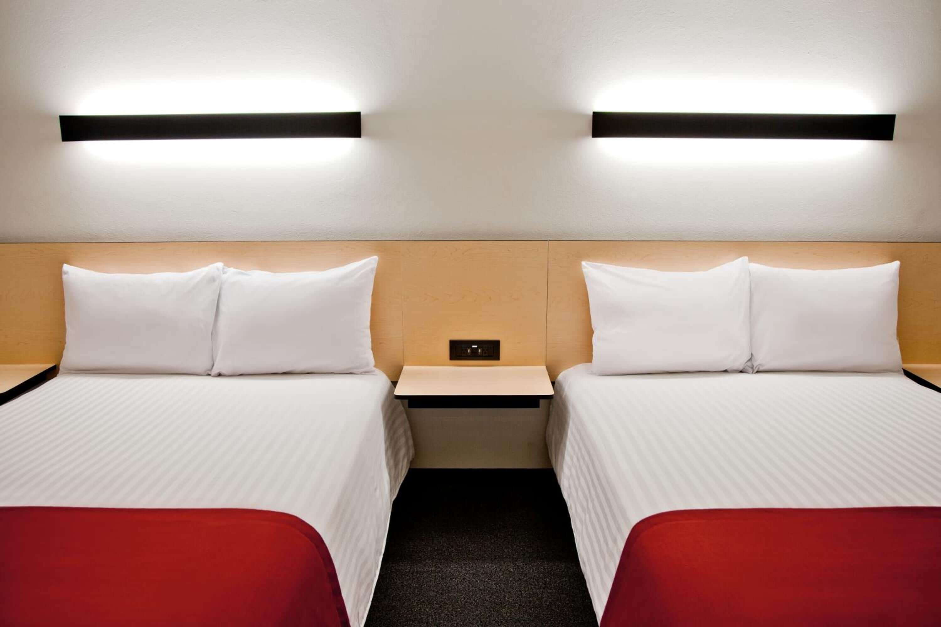 Ett hotellrum med två sängar, båda med vita sängkläder och röda prydnadskuddar. Ovanför sängarna finns väggmonterade lampor.