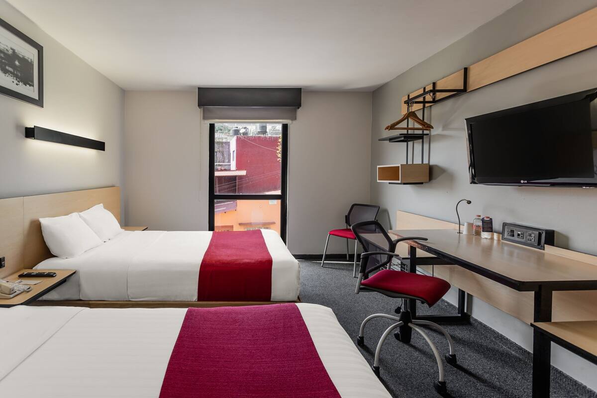 A hotel room with two beds, a desk, a chair, a TV, and a window with a view of buildings.