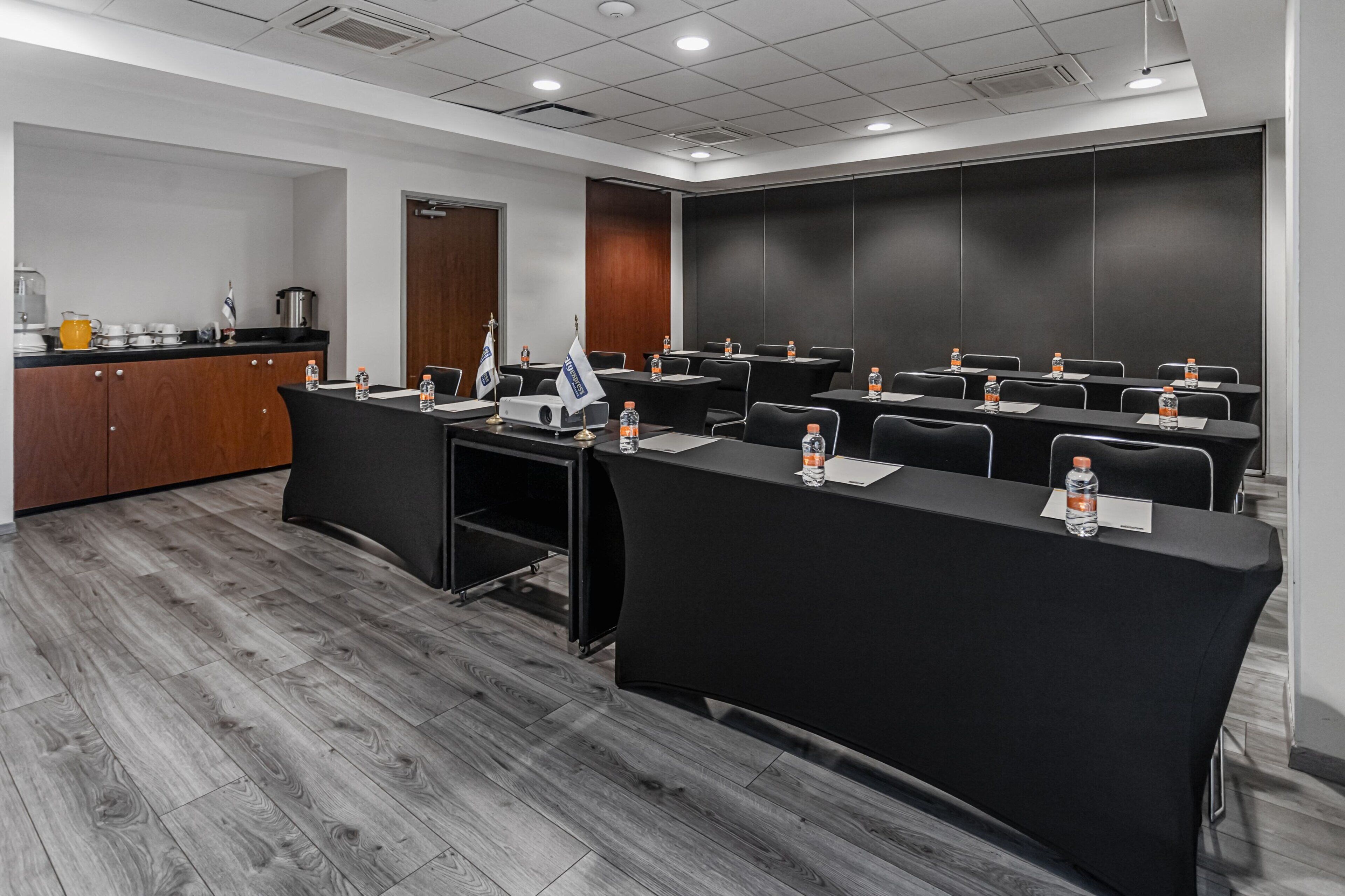 A conference room with a long table, chairs, and a kitchen area with a coffee machine and water bottles.