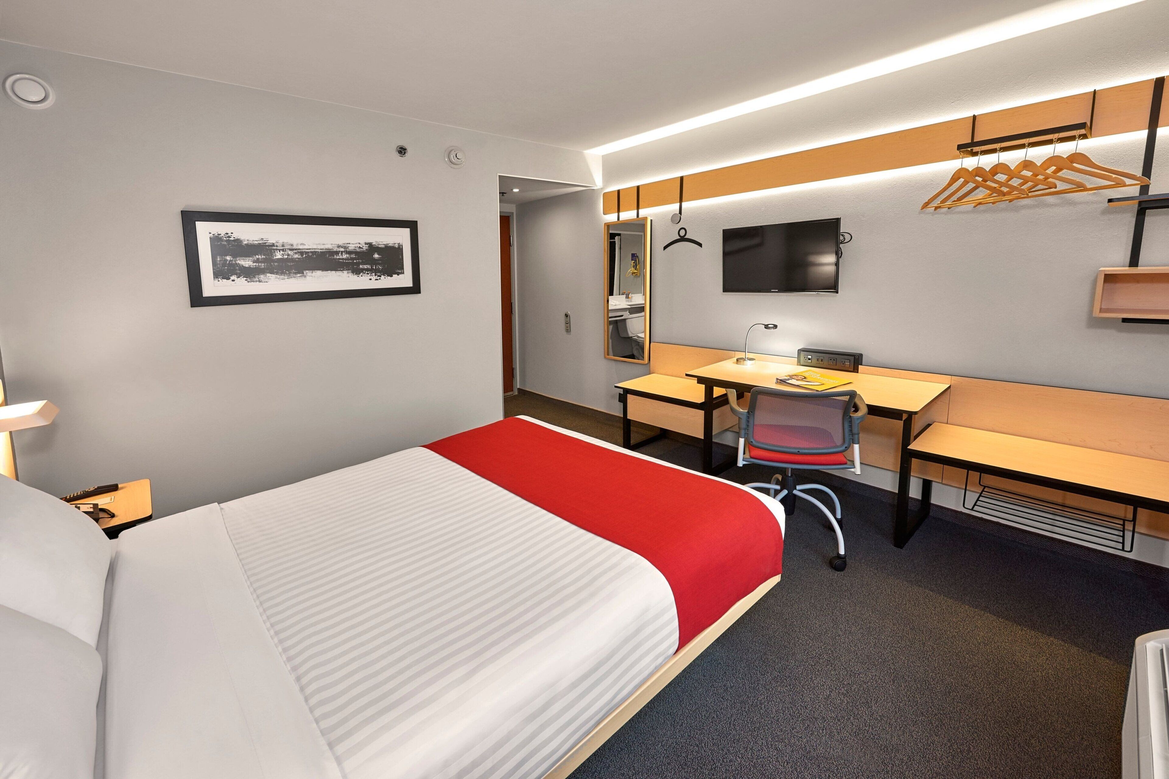 A hotel room with a bed, a desk, a chair, a mirror, a TV, and a coat rack.