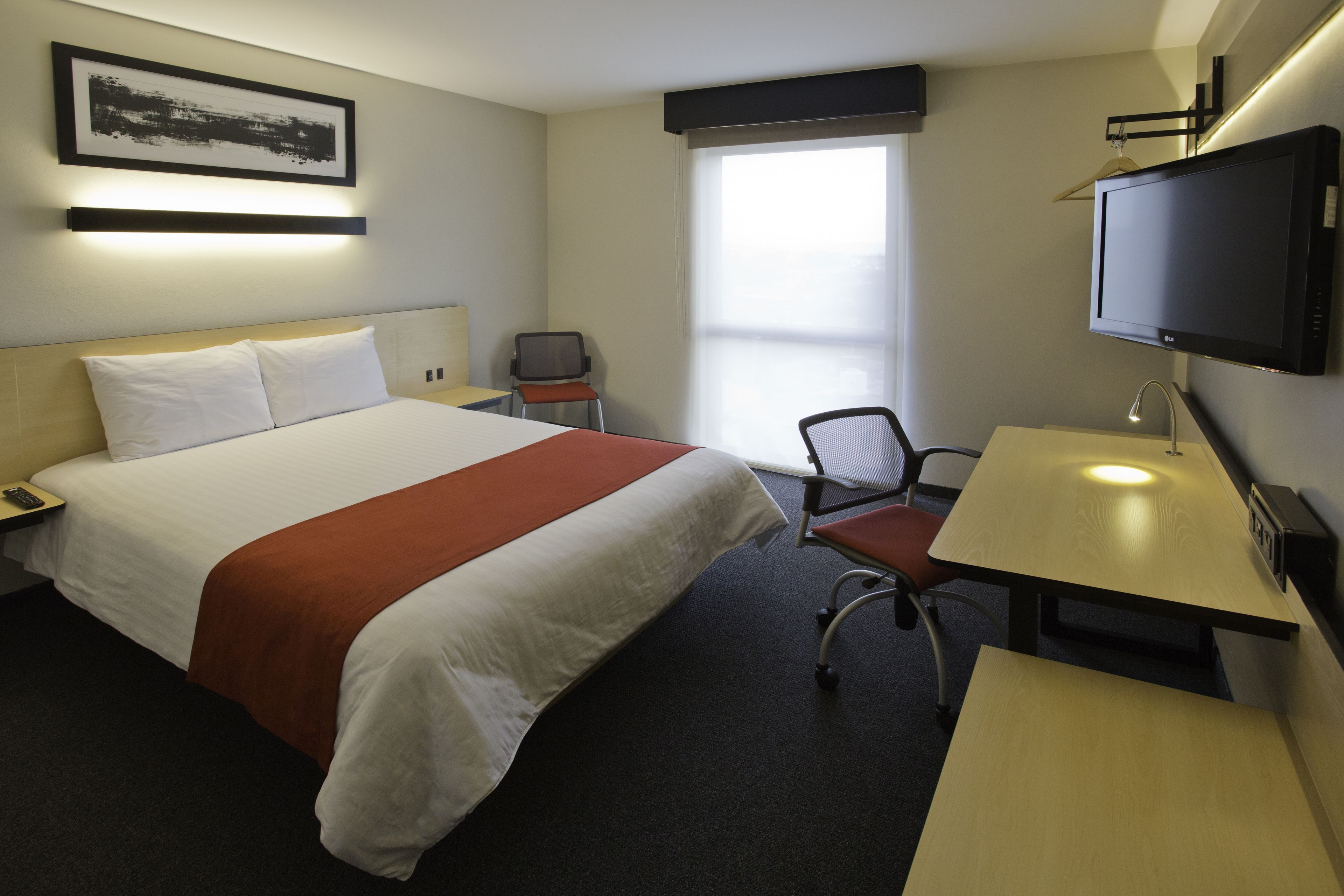 A hotel room with a bed, a desk, a chair, a television, and a window.