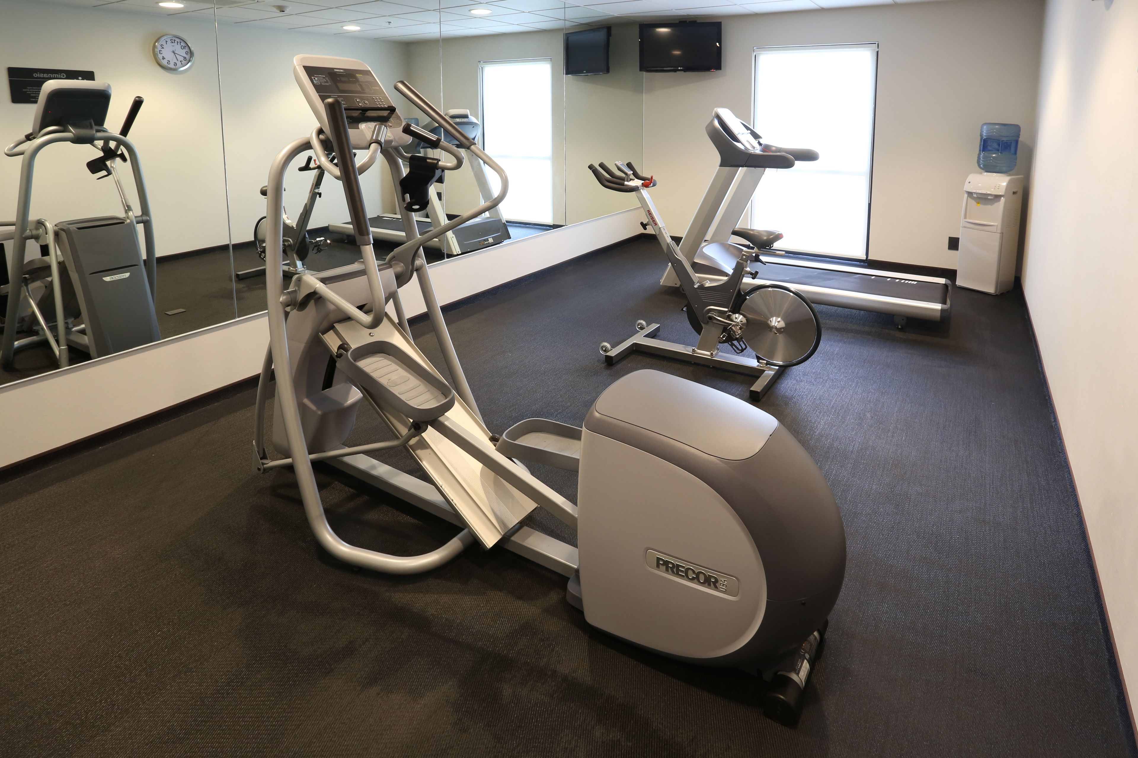 Un gimnasio con máquinas de cardio, un dispensador de agua y un reloj en la pared.