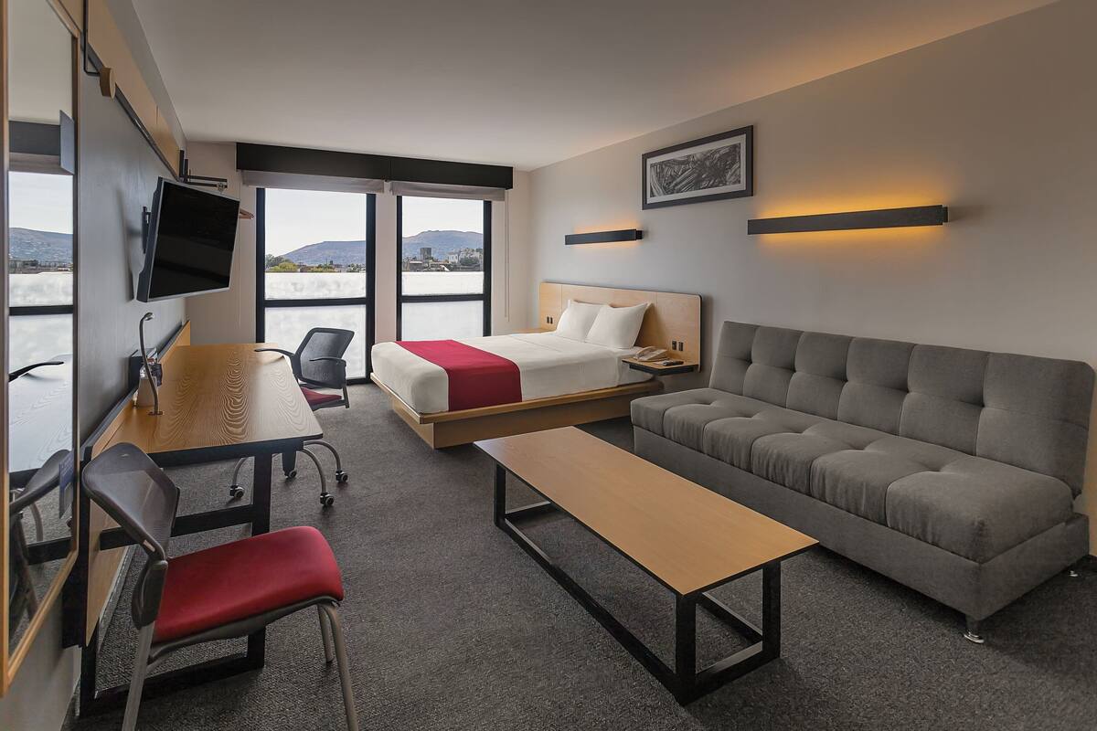 A hotel room with a bed, a sofa, a desk with a chair, a TV mounted on the wall, and a large window offering a view of the city.