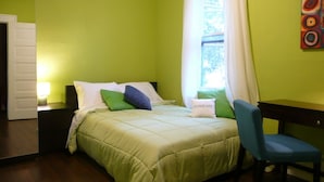 Een slaapkamer met een bed, kussens, een stoel, een nachtkastje en een raam met gordijnen.