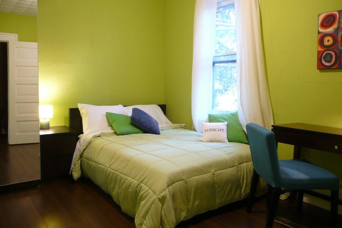 Een slaapkamer met een bed, kussens, een stoel, een nachtkastje en een raam met gordijnen.