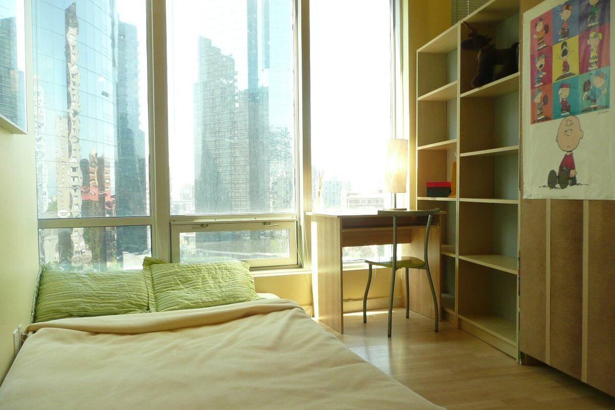 A bedroom with a bed, a desk, and a chair. There is a window with a city view.