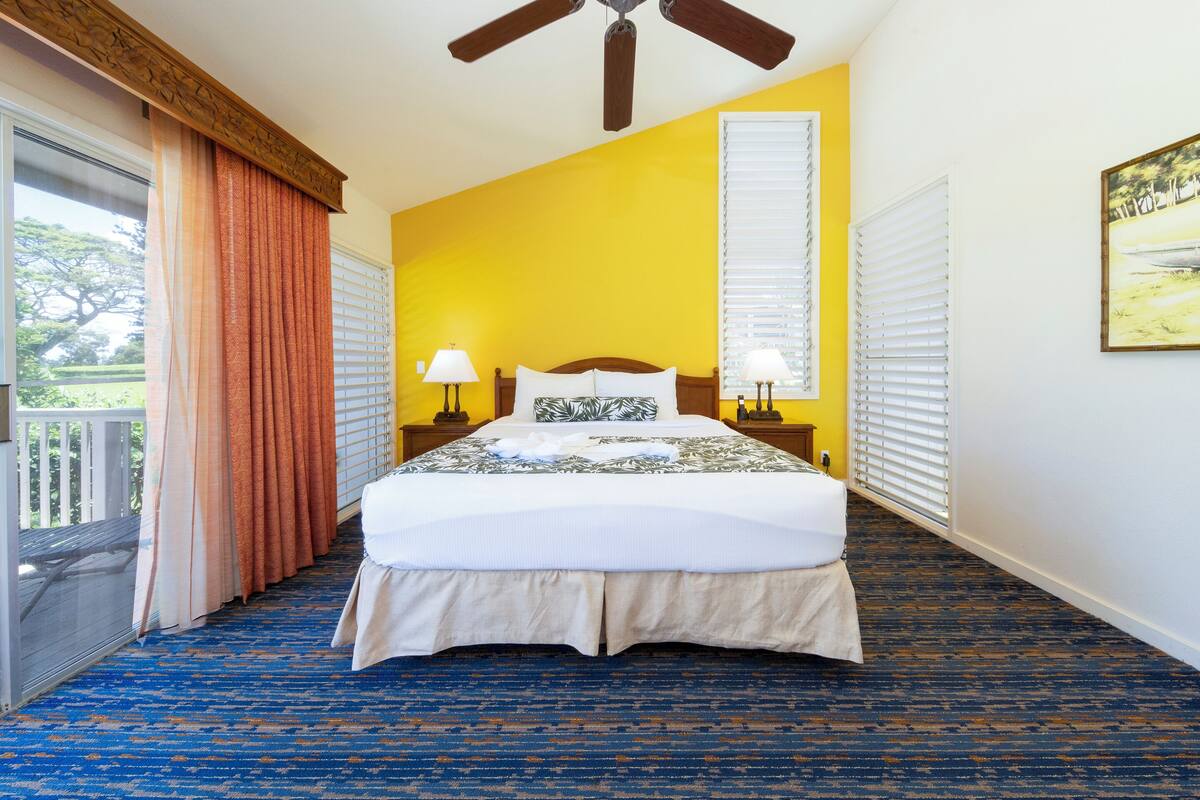 A hotel room with a large bed, two bedside lamps, a ceiling fan, and a view of the outdoors.