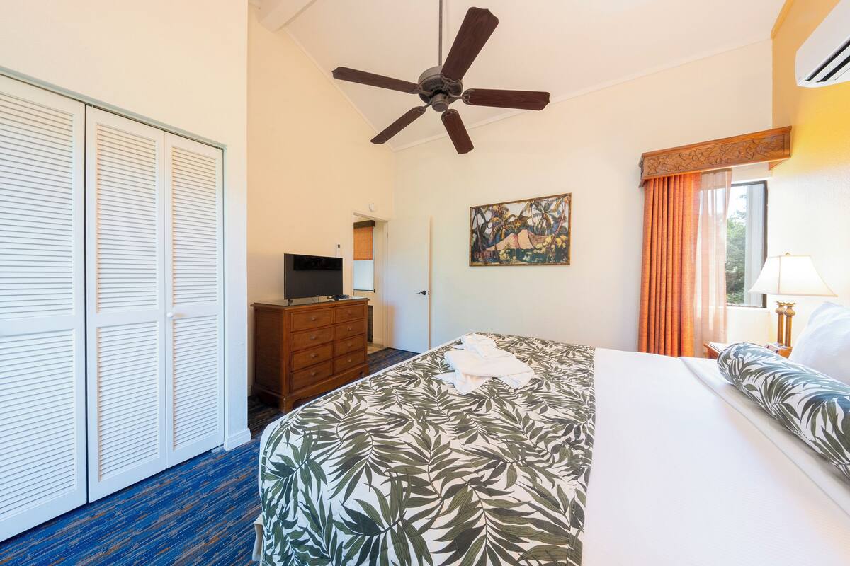 A hotel room with a bed, a ceiling fan, a television, a painting, and a window with curtains.