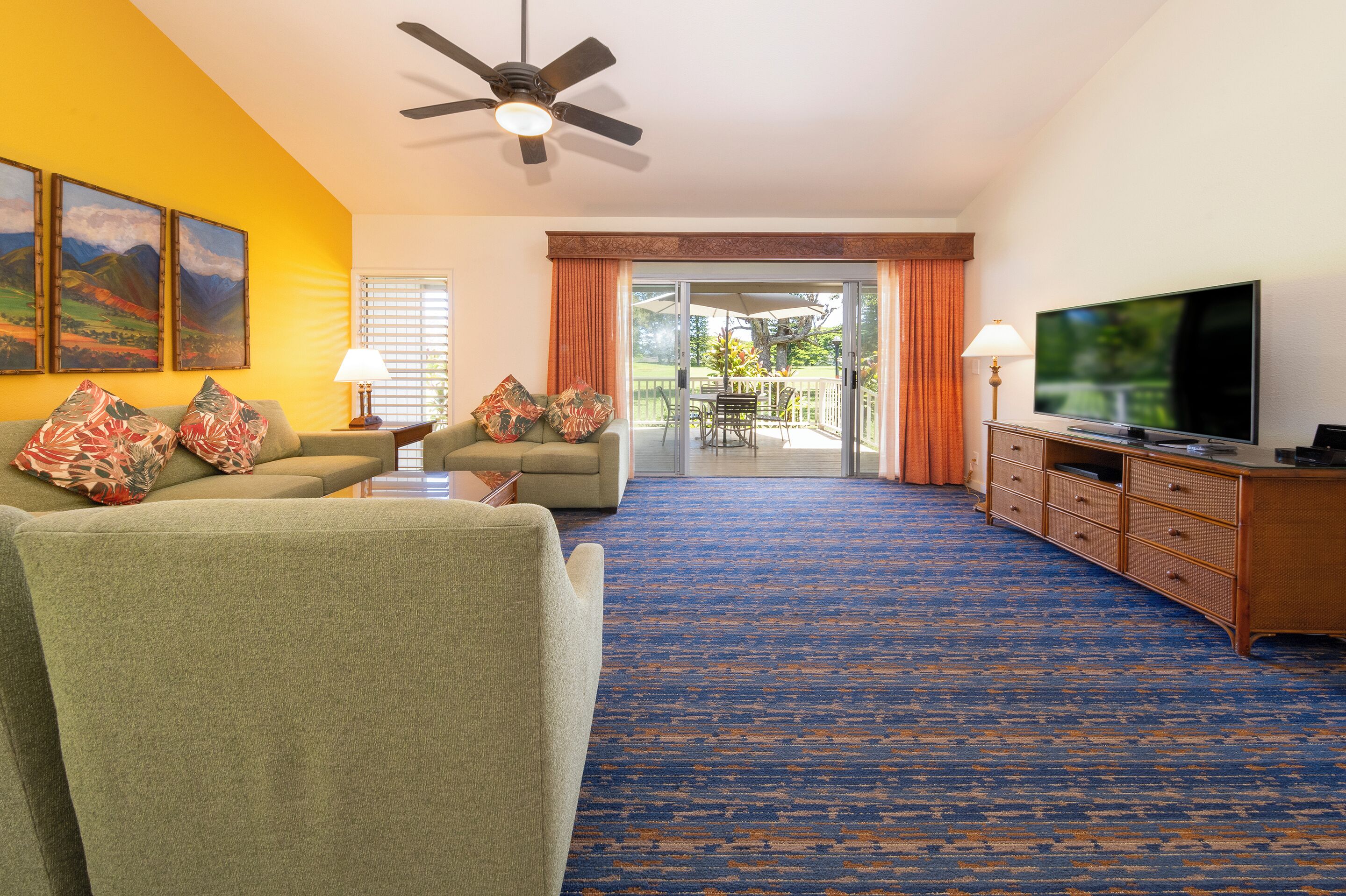 A hotel room with a sofa, armchairs, a television, a ceiling fan, and a view of the outdoor patio.
