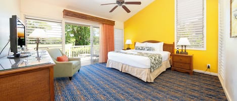 A hotel room with a bed, a desk, a chair, a ceiling fan, and a balcony with a view of trees.
