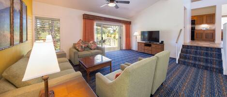 A hotel room with a living area featuring a sofa, a coffee table, a TV, and a ceiling fan. There is a sliding glass door leading to a balcony.