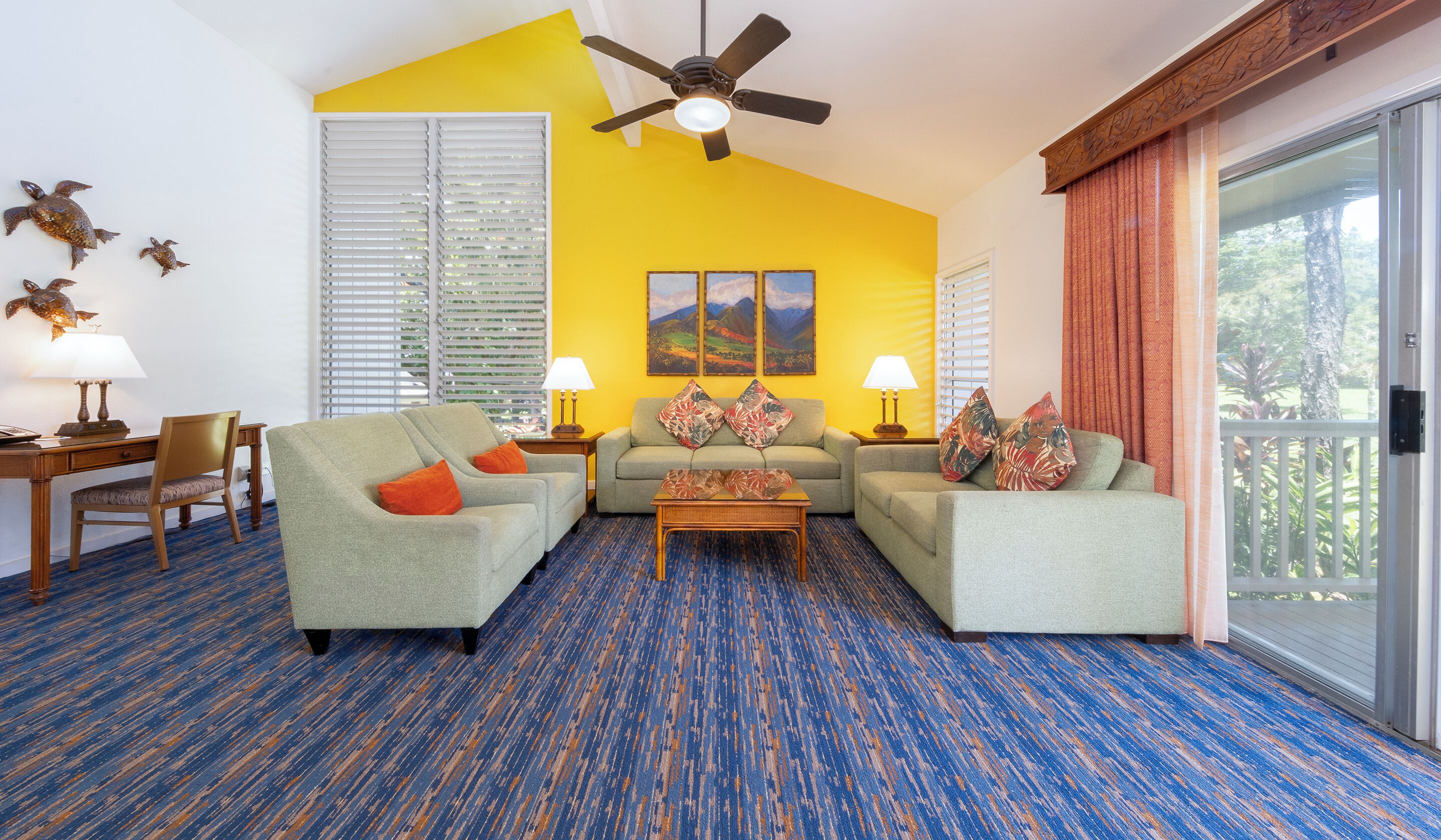 A living room with a blue and orange carpet, two grey sofas with orange cushions, a wooden coffee table, and a ceiling fan.