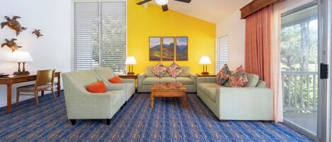 A living room with a blue and orange carpet, two grey sofas with orange cushions, a wooden coffee table, and a ceiling fan.