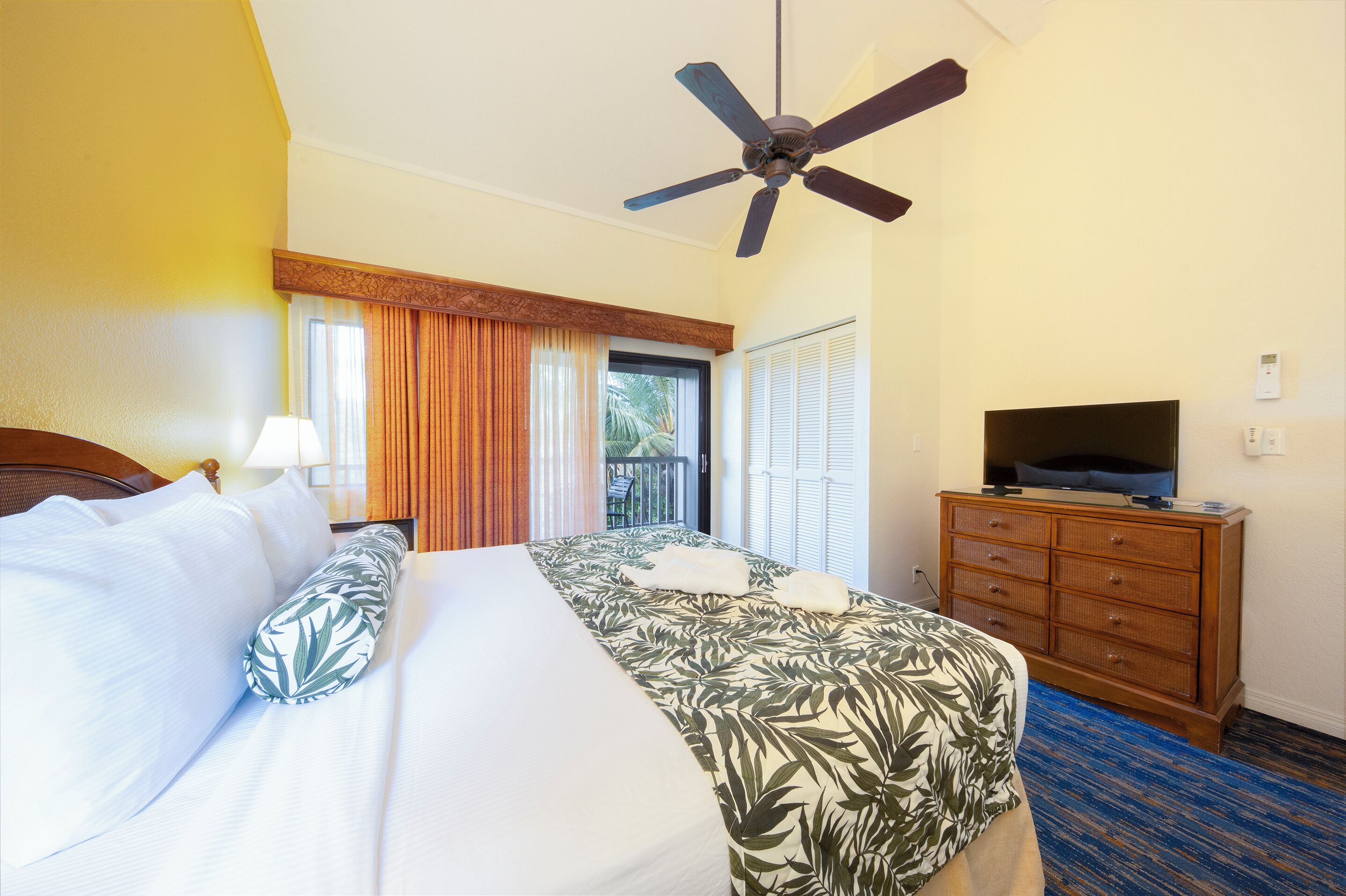 A hotel room with a bed, a television on a wooden dresser, a ceiling fan, and a window with curtains.