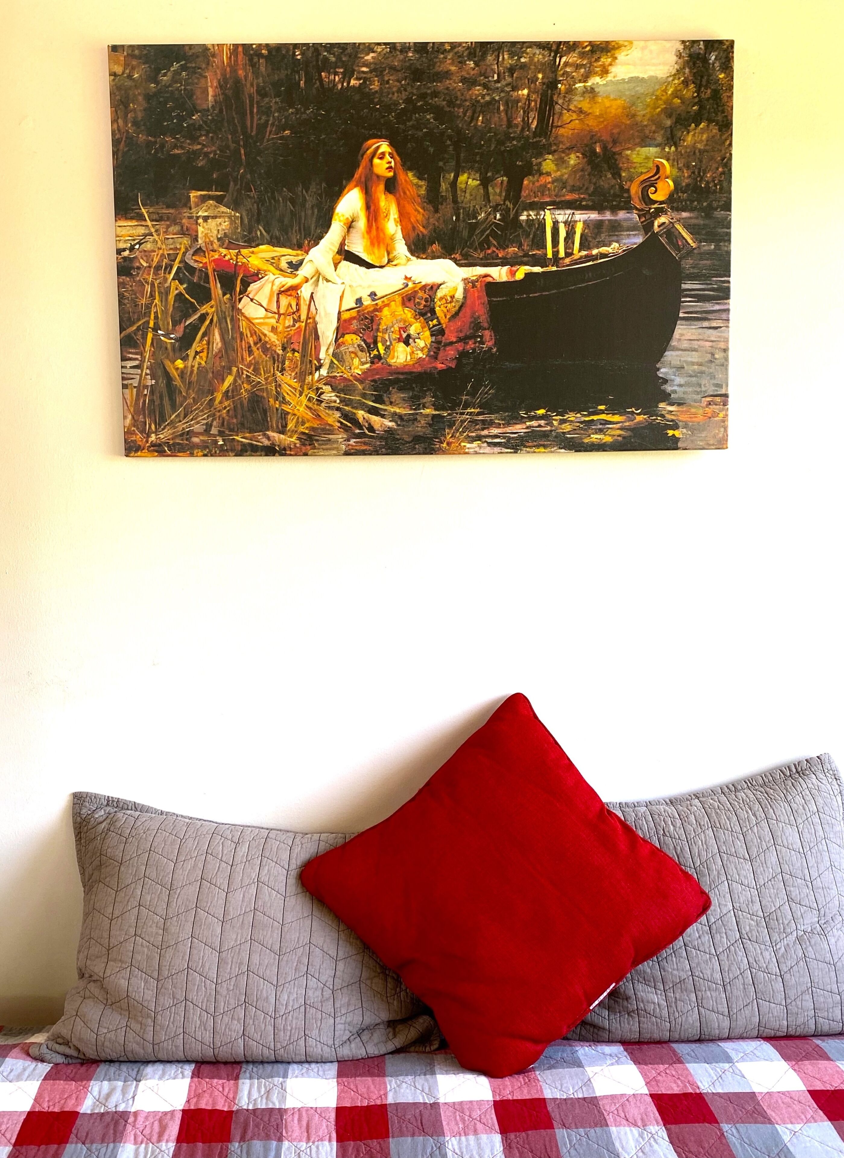 Un tableau représentant une femme au bord d’une rivière, un coussin rouge et une couverture à carreaux.