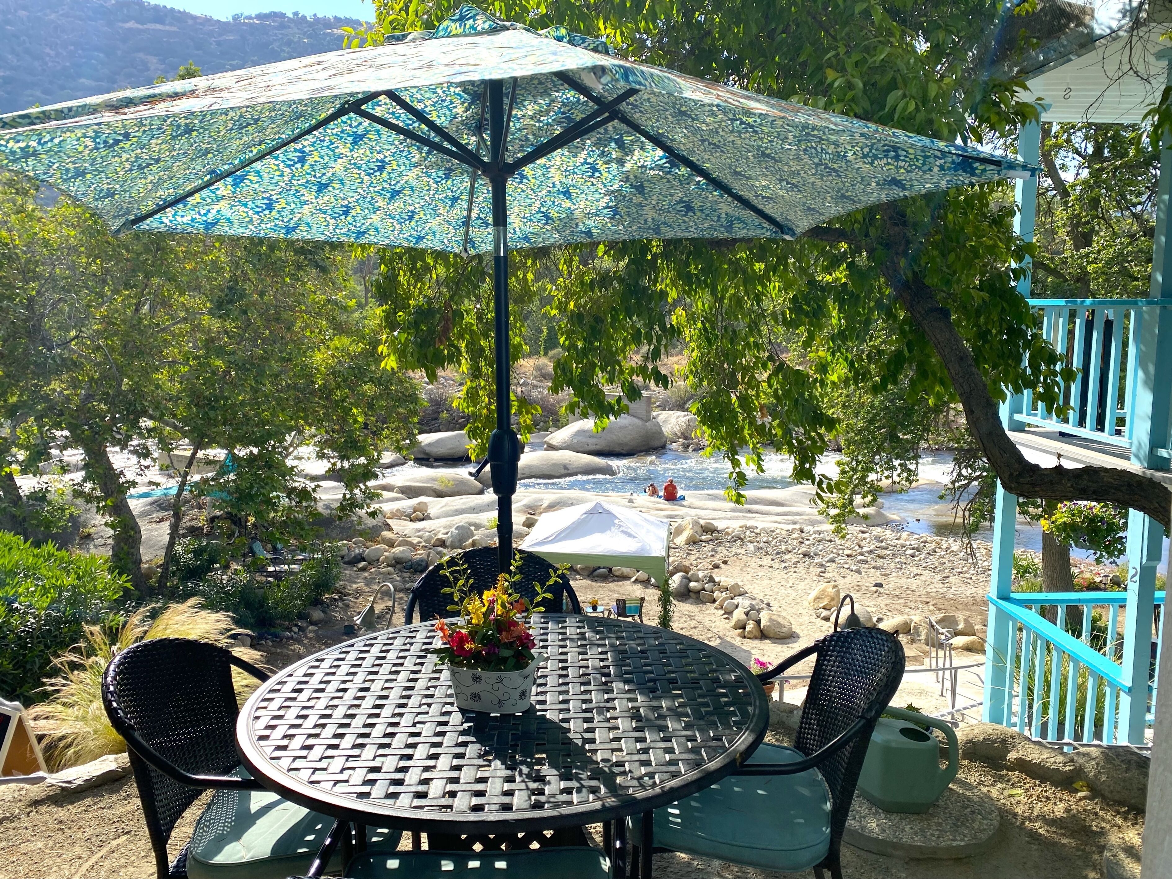 An outdoor seating area with a table and chairs, an umbrella, and a view of a river and rocks.