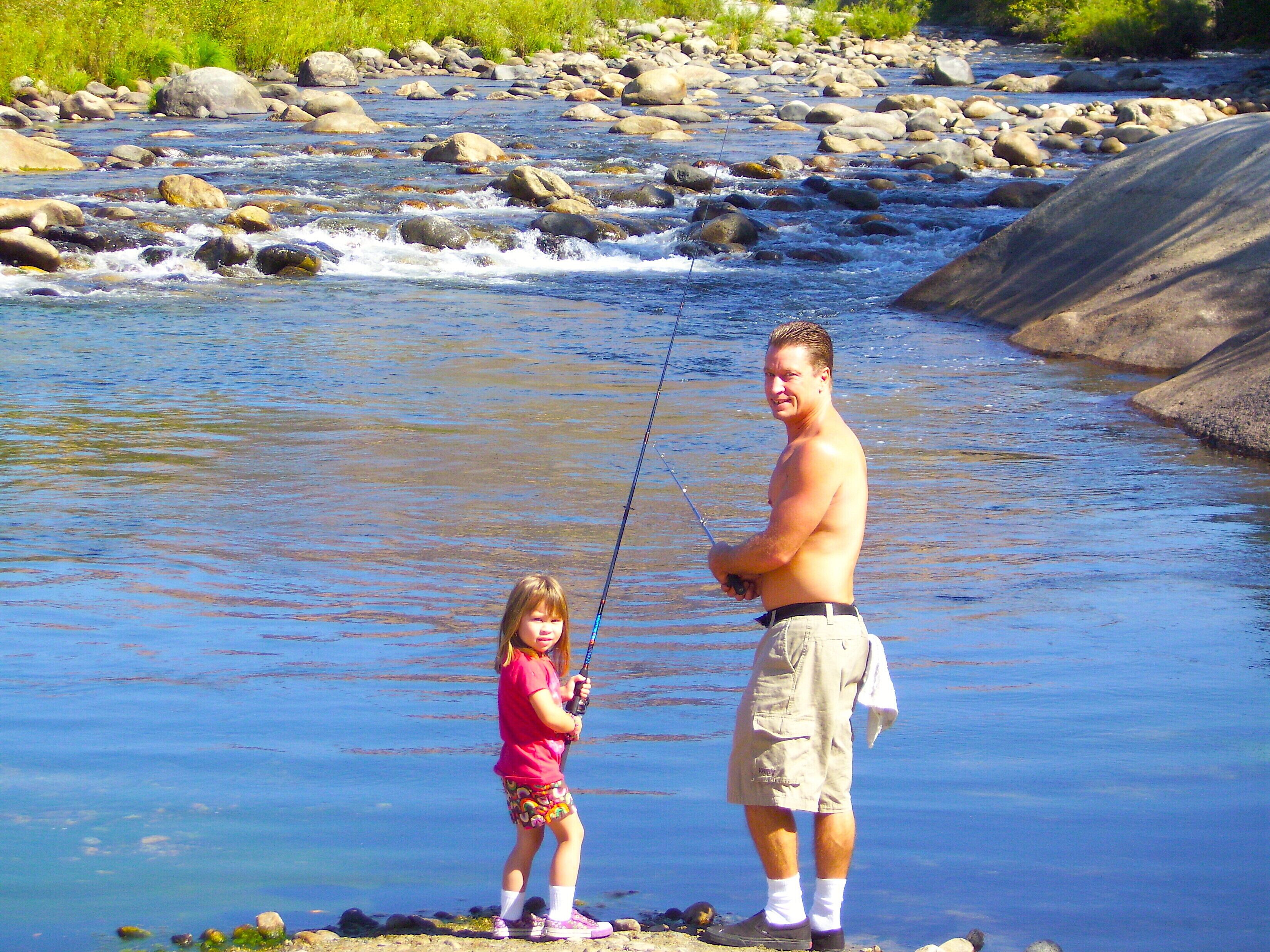A man and a child fishing by a river with rocks and greenery in the background.