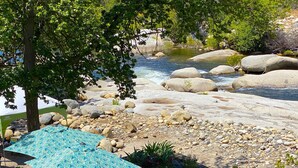 A riverside outdoor seating area with a table and chairs under a blue umbrella, surrounded by rocks and greenery.