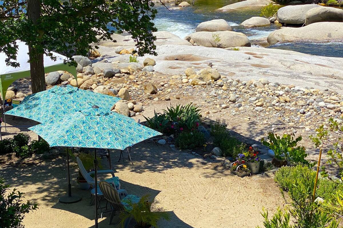 A riverside outdoor seating area with a table and chairs under a blue umbrella, surrounded by rocks and greenery.