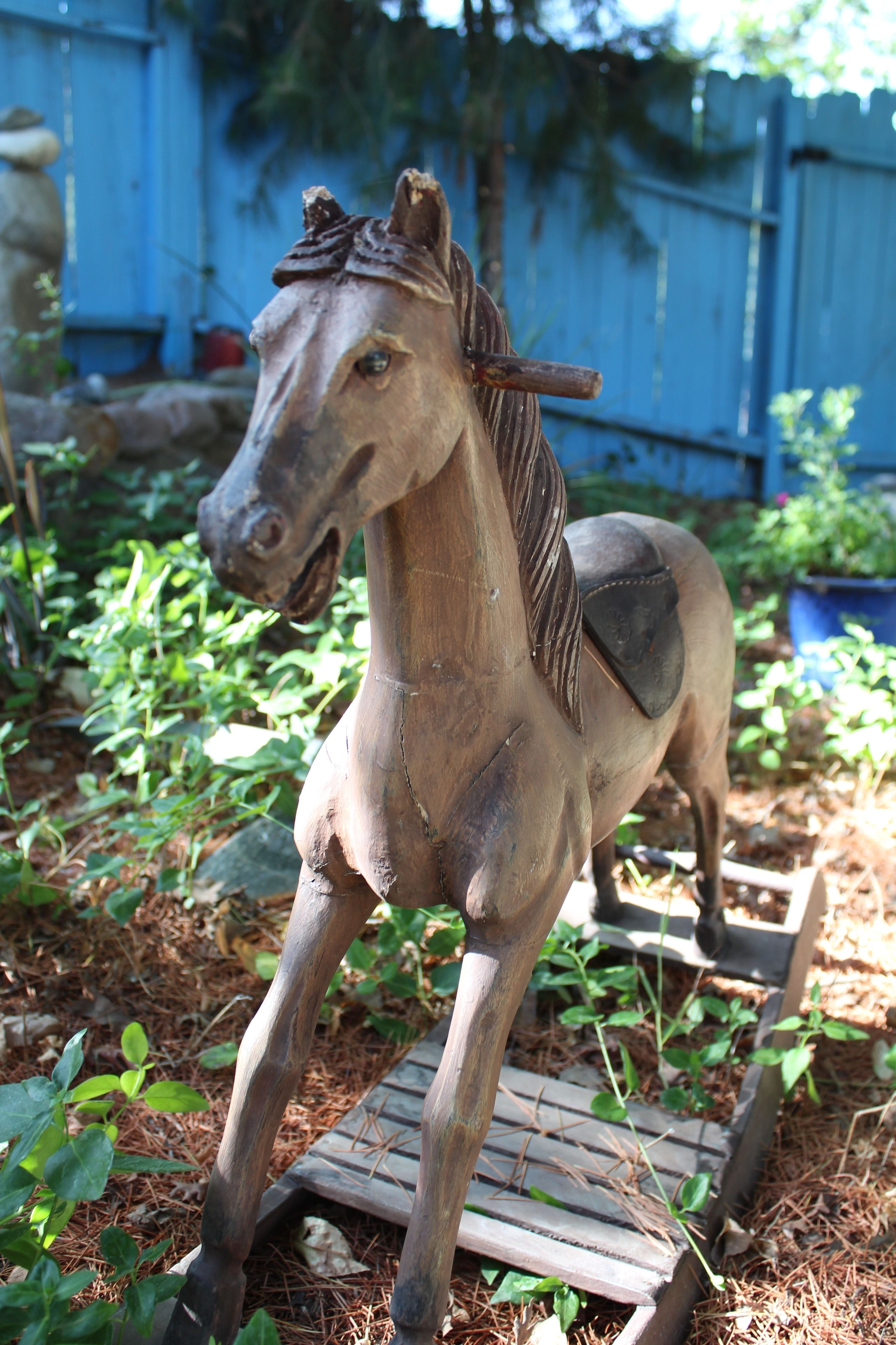 A wooden horse sculpture in a garden with a blue fence and greenery in the background.