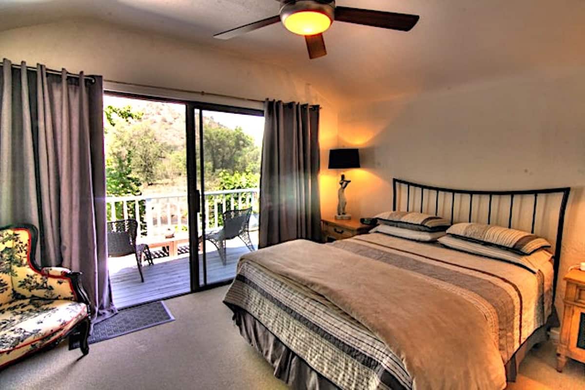 A bedroom with a bed, a ceiling fan, a nightstand, a lamp, a chair, and a view of the outdoors.