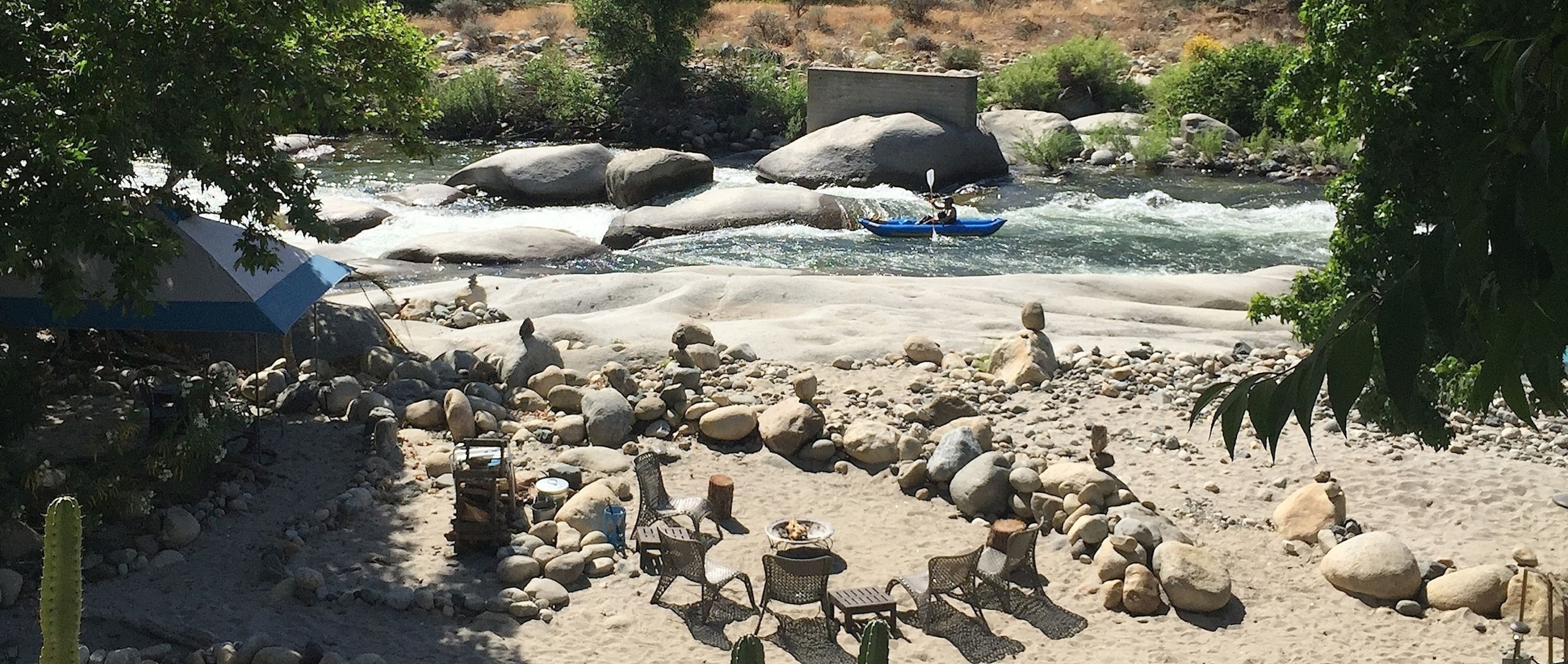 A riverside campsite with a kayaker, rocks, and camping chairs.