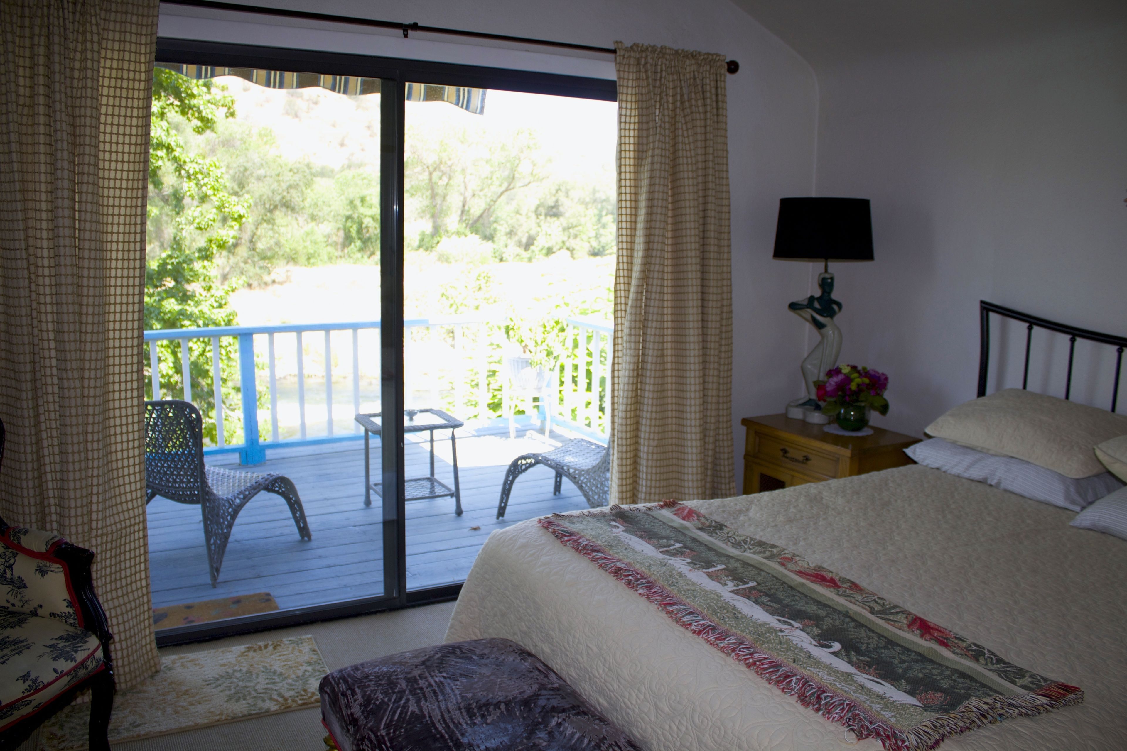 Une chambre avec un grand lit, une table de chevet avec une lampe, une chaise et une vue sur un balcon aménagé avec du mobilier d’extérieur.