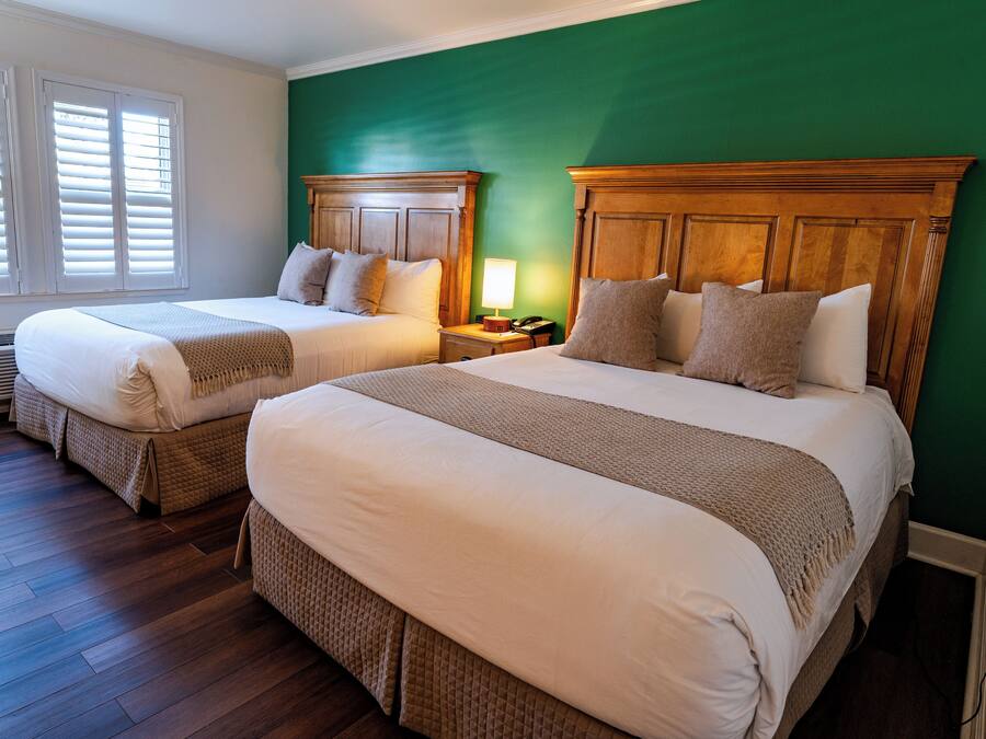 A hotel room with two beds, wooden headboards, a green accent wall, and a window with blinds.