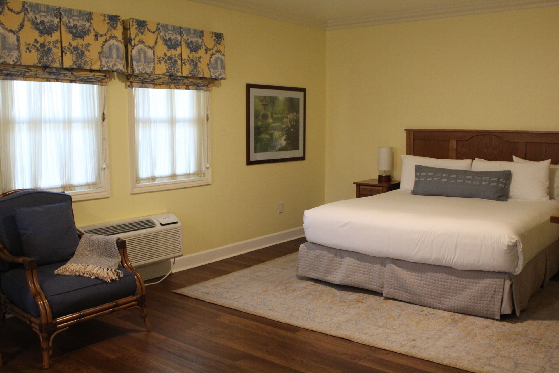 A bedroom with a large bed, a chair, a framed picture, and a window with curtains.