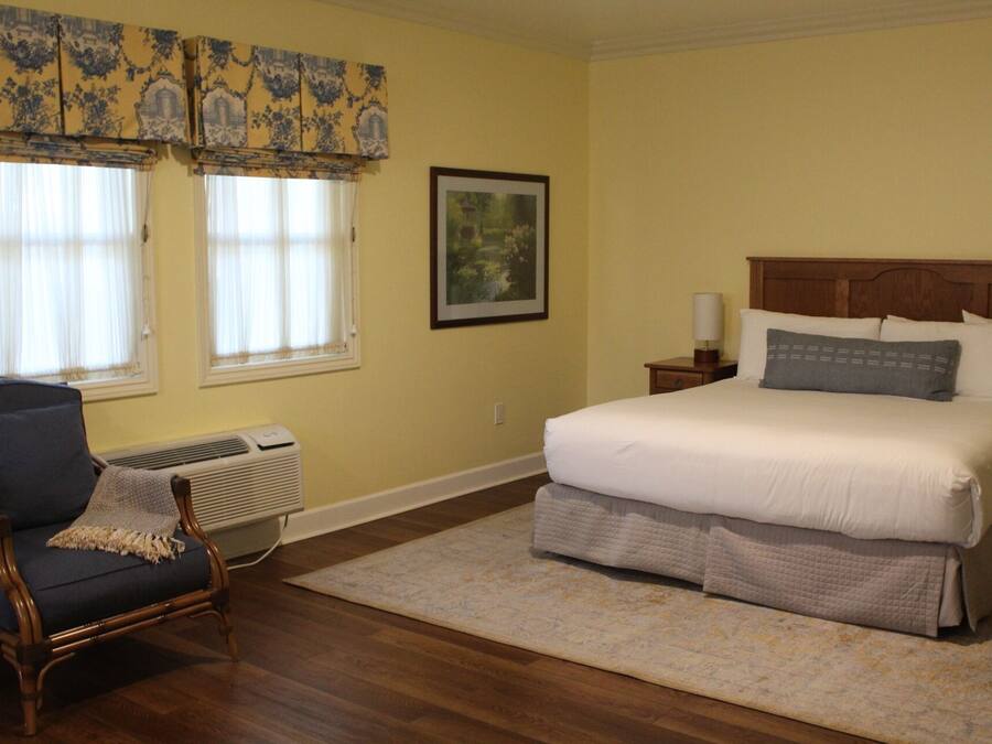 A bedroom with a large bed, a chair, a framed picture, and a window with curtains.