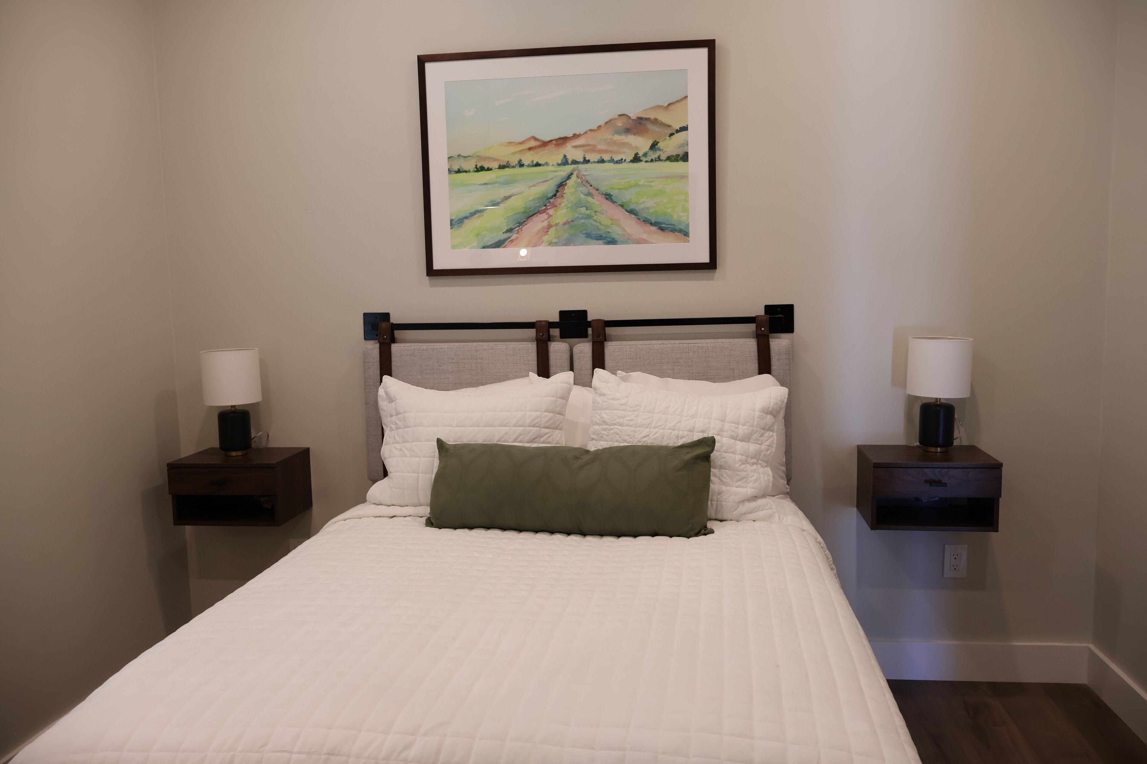 A neatly made bed with white bedding and a green pillow, flanked by two bedside tables with lamps, in a room with a framed landscape painting above the bed.